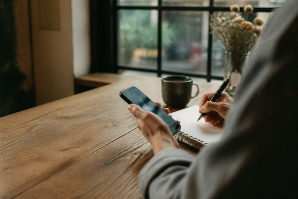 Anonymous adult drafting a thoughtful note beside a phone at a cafe table.